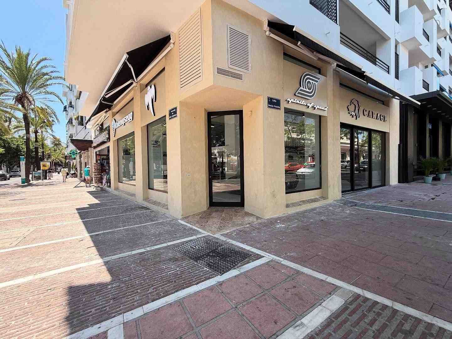 Esquina soleada con una tienda de color beige, ventanas de cristal y una acera bordeada de palmeras.