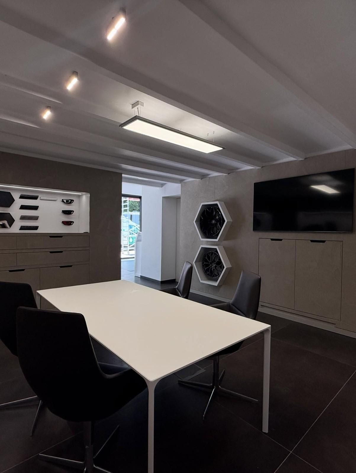 Sala de reuniones de oficina moderna con mesa blanca, sillas negras, televisor montado en la pared y estantes hexagonales.