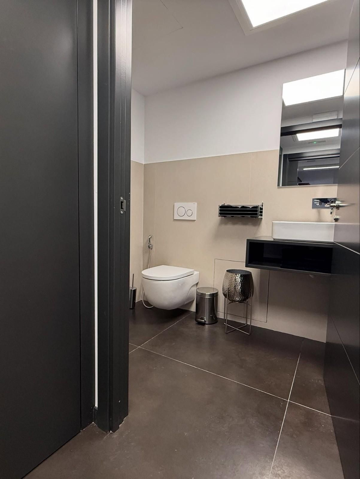 Baño público con lavabo, espejo, secador de manos y papelera, visto a través de una puerta oscura.
