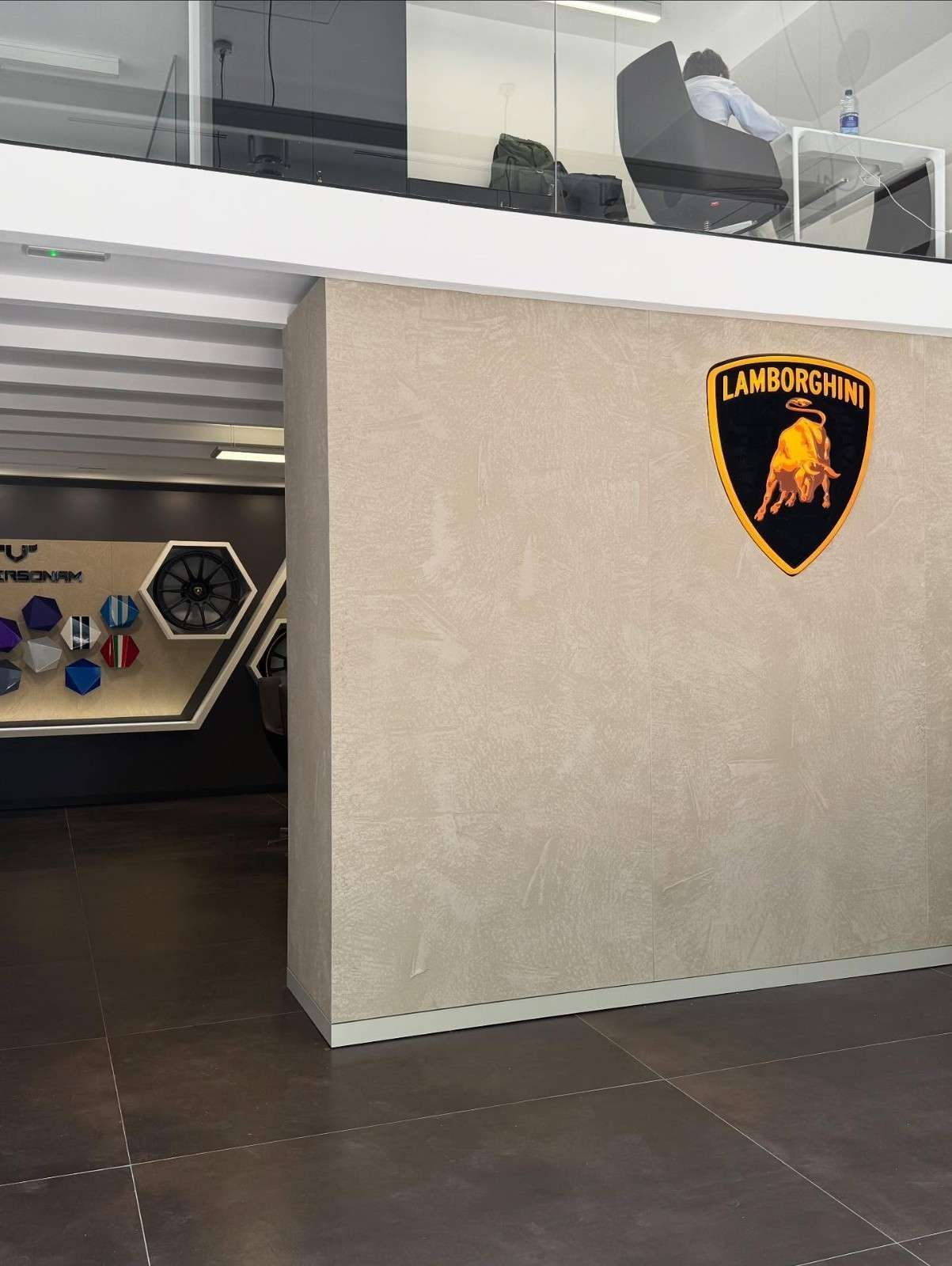 Entrada a la sala de exposición de Lamborghini con logotipo del escudo, barandilla de cristal y suelo de baldosas oscuras.