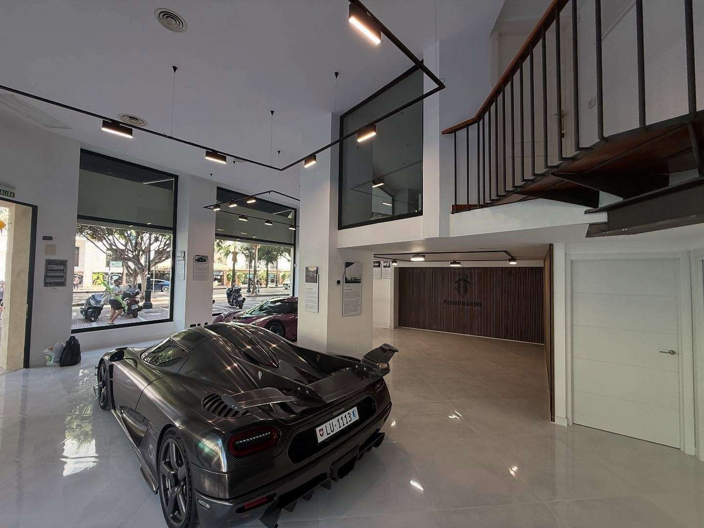 Coche deportivo negro aparcado en una luminosa y moderna sala de exposición con techo alto y escalera de balcón.