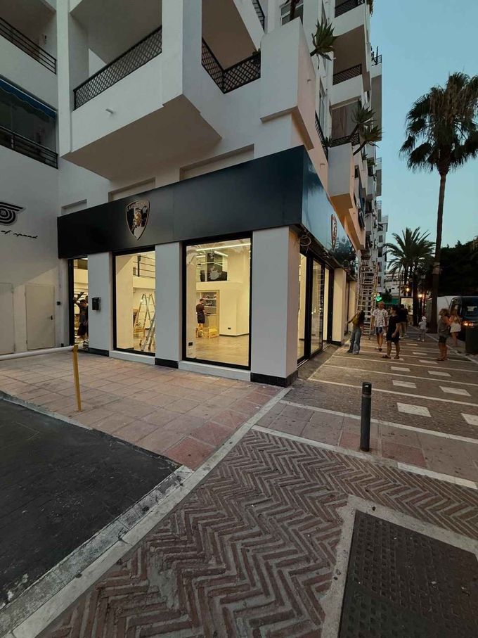 Esquina de la calle con una tienda moderna debajo de los apartamentos, ventanas iluminadas y un paseo bordeado de palmeras al atardecer.