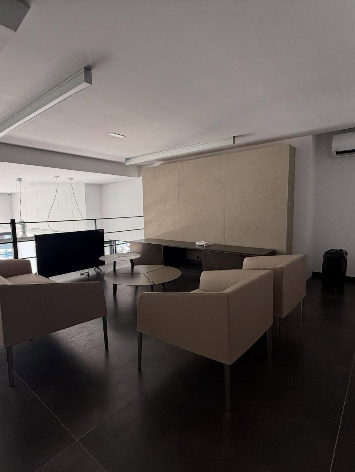 Sala de oficina moderna con sillones beige, un escritorio con monitor y grandes ventanales en un interior minimalista.
