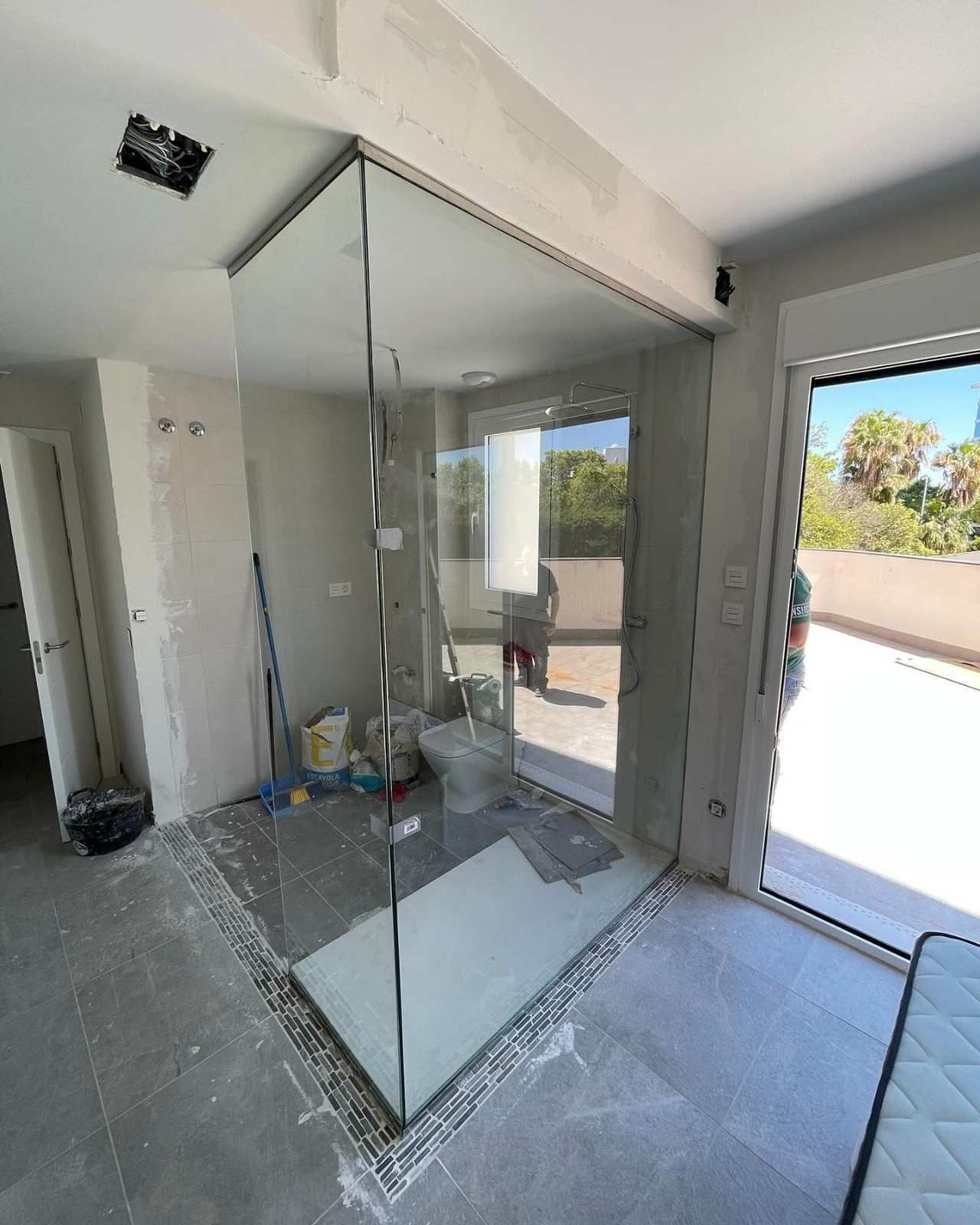 Mampara de ducha moderna de cristal en un baño sin terminar con suelo de baldosas grises.