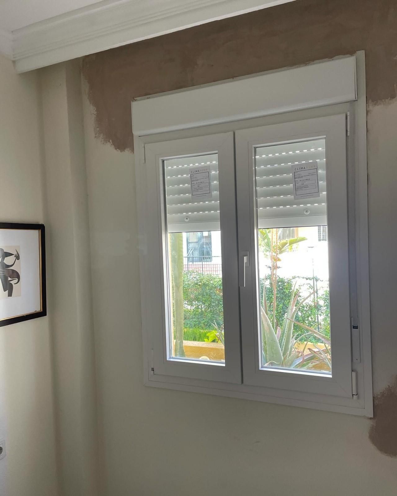 Ventana blanca con contraventanas y plantas en el exterior, enmarcada por paredes parcialmente pintadas de color beige.