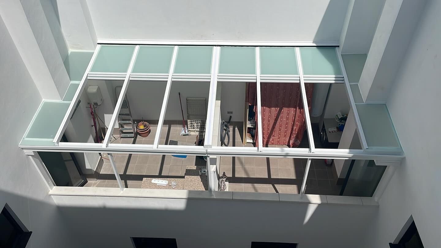 Balcón con barandilla blanca, una persona sentada y una manta roja colgada en el interior, detrás de un cristal.