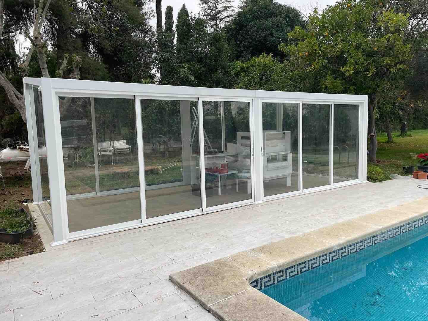 Pabellón de piscina blanco acristalado junto a una piscina en un entorno ajardinado.