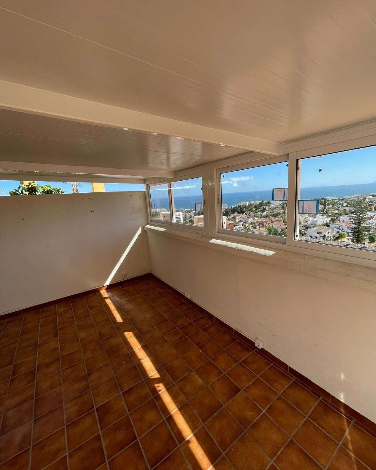 Terraza vacía con baldosas, paredes blancas y ventanas abiertas con vistas a la ciudad y al mar.