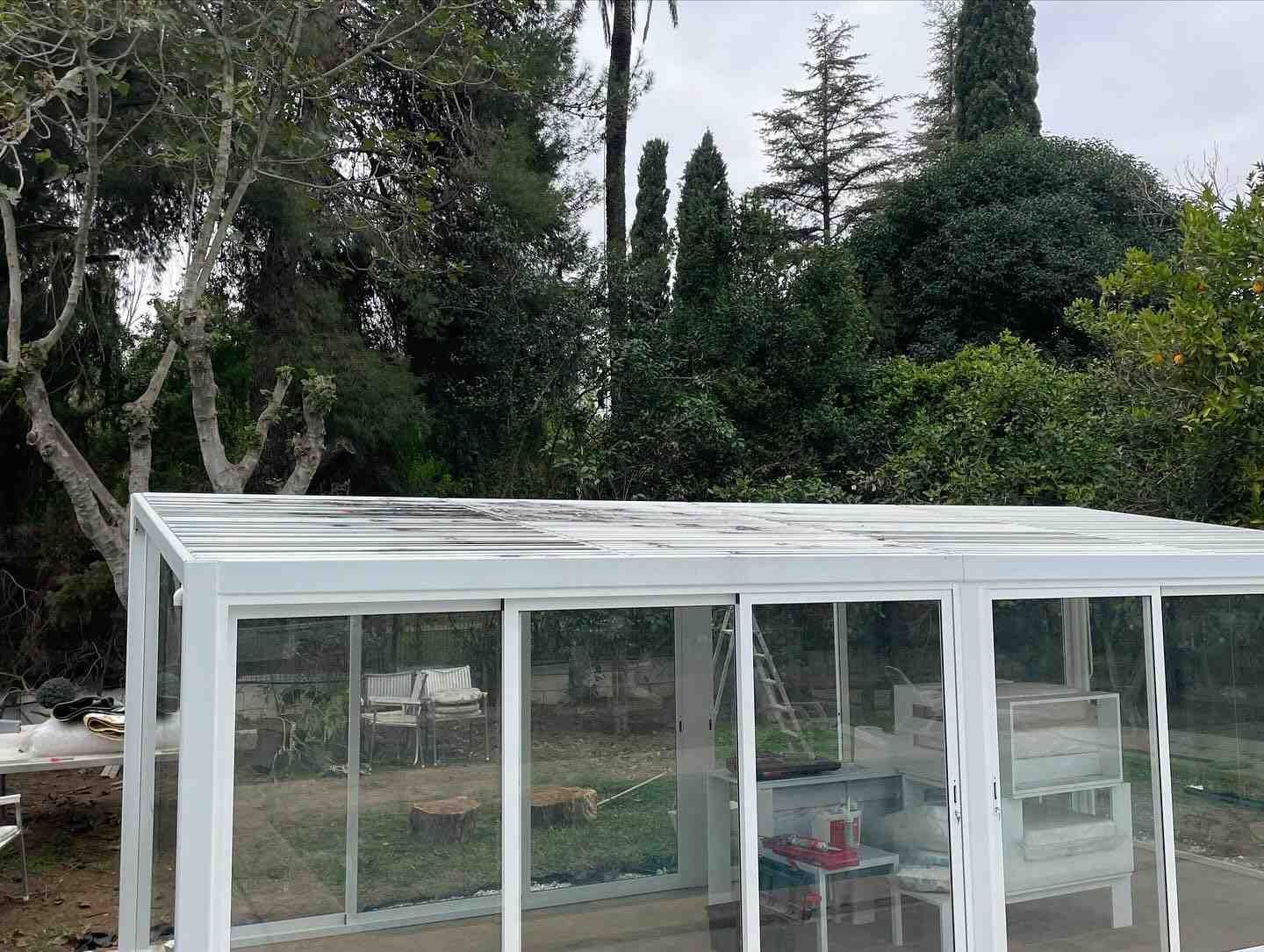 Invernadero de cristal blanco en un jardín, con árboles altos y vegetación densa al fondo.
