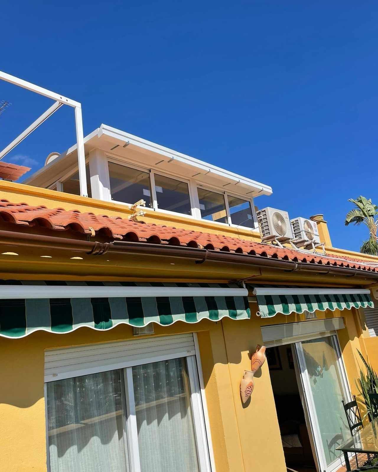 Casa amarilla con techo de terracota y toldo verde bajo un cielo azul brillante.