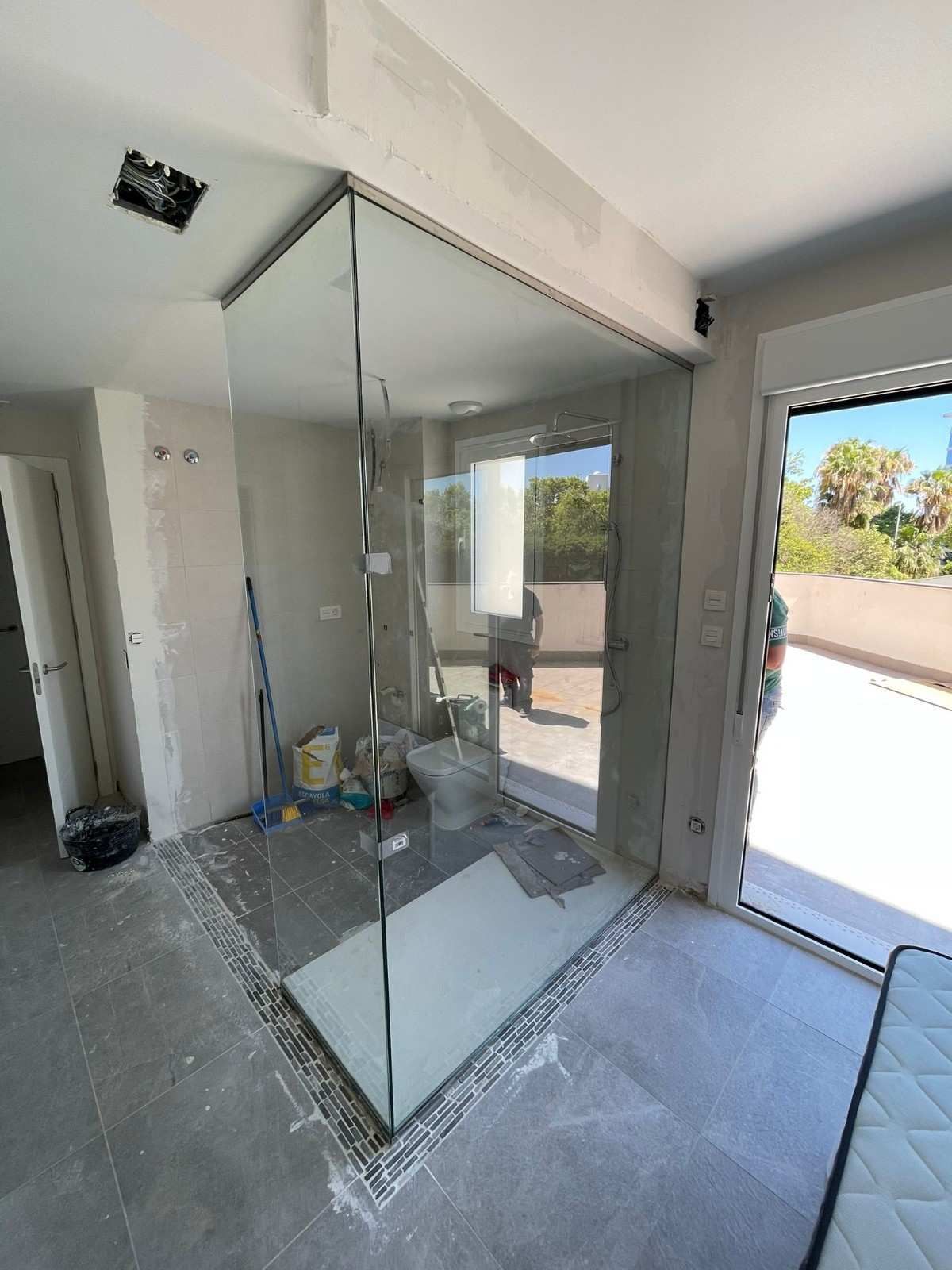 Mampara de ducha de cristal en construcción en un baño luminoso, con suelo de baldosas y puerta abierta al exterior.