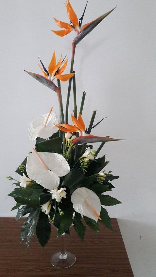 Grande composition florale avec des oiseaux de paradis orange et des fleurs blanches dans un vase en verre sur une table.