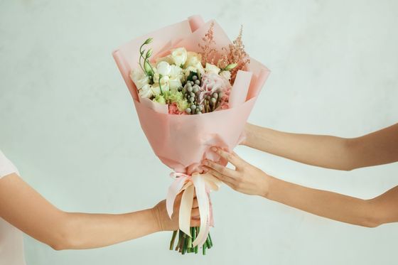 Des mains présentent un bouquet de fleurs enveloppé de rose sur un fond clair.