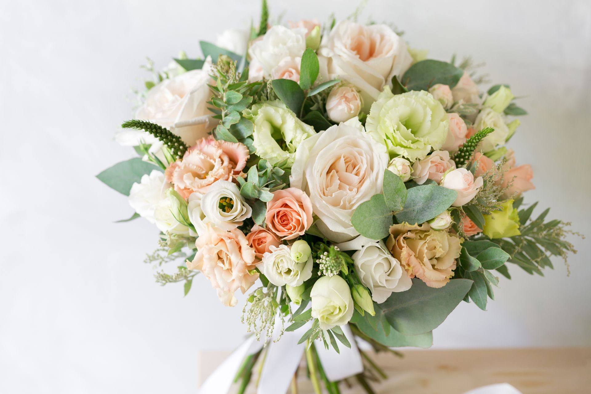 Bouquet de roses crème et pêche avec feuillage et ruban blanc sur fond clair.