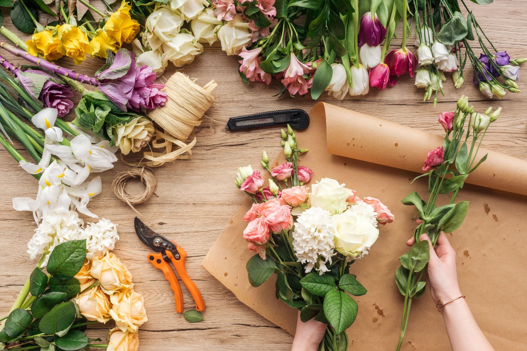 Des bouquets de fleurs et des outils sont posés sur une table en bois, des mains arrangeant des fleurs roses et blanches.