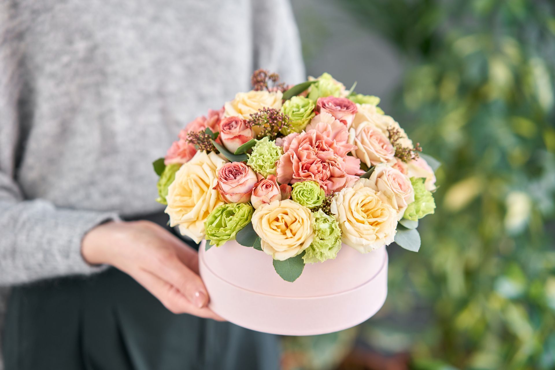 Des roses pastel dans une boîte à chapeau rose tenue par une personne en pull gris.