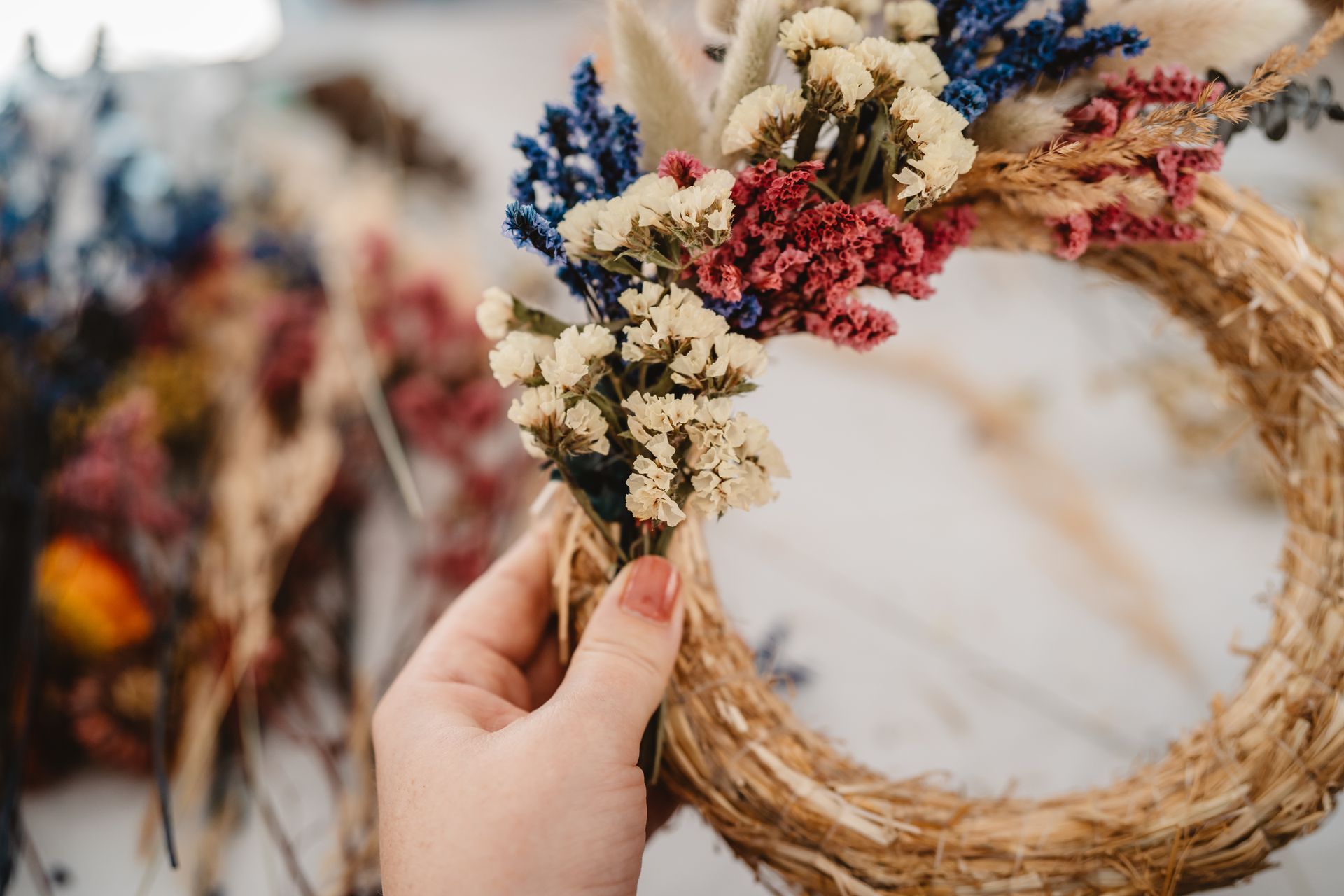 Main tenant une couronne de fleurs séchées et de paille tressée avec des accents crème, bleus et roses.