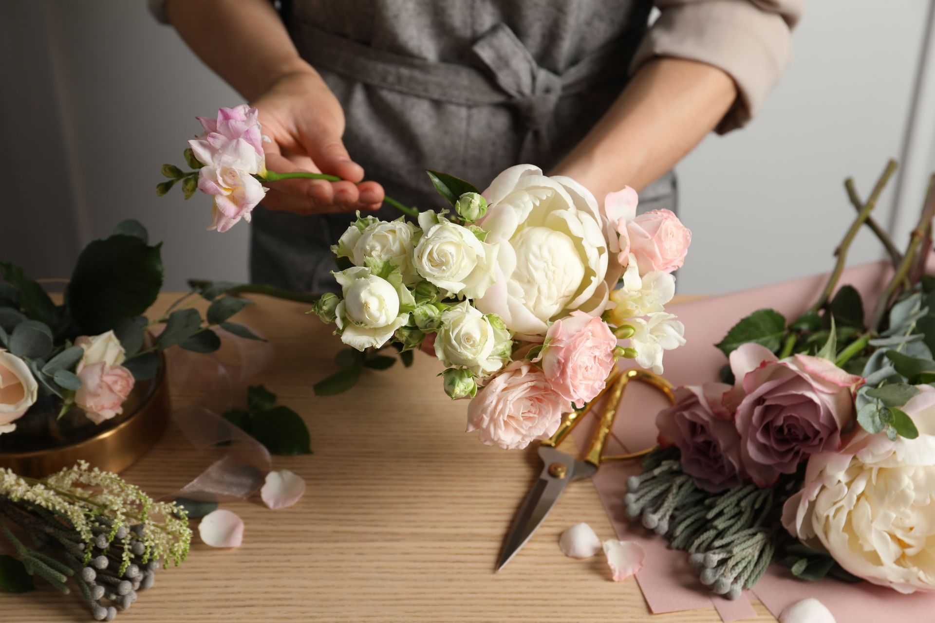Des mains composent un bouquet de fleurs pastel sur une table en bois, à l'aide de ciseaux et de fleurs éparpillées.