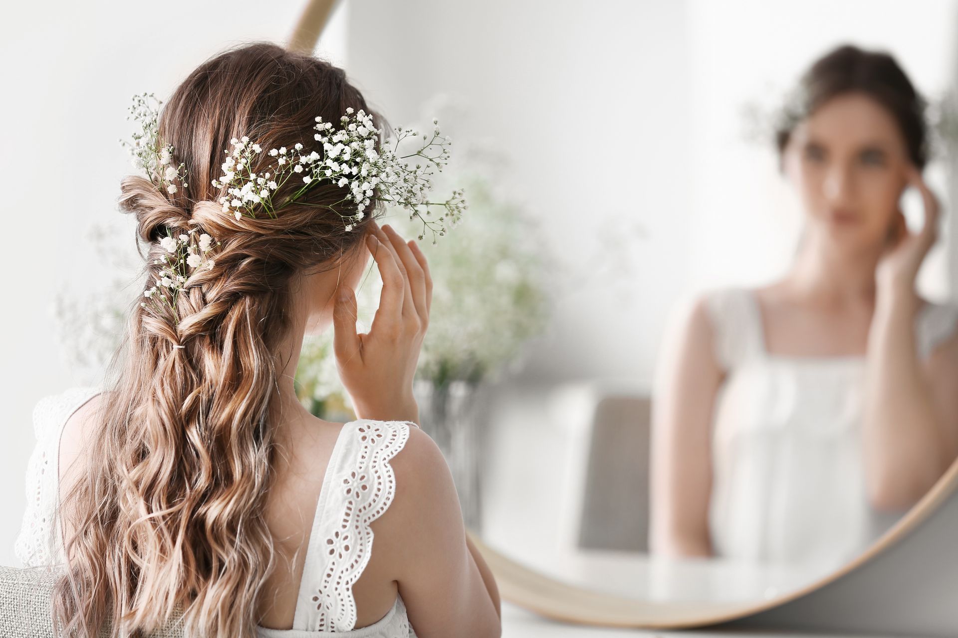 Une femme aux cheveux tressés et parée de fleurs blanches se regarde dans un miroir dans une pièce lumineuse.
