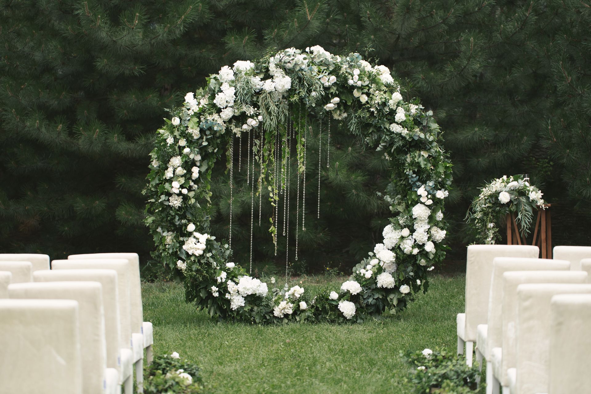Arche de mariage florale blanche sur une allée gazonnée, bordée de rangées de chaises blanches en extérieur.