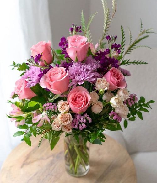 Bouquet de roses roses et de fleurs sauvages violettes dans un vase en verre, posé sur une table ronde en bois.