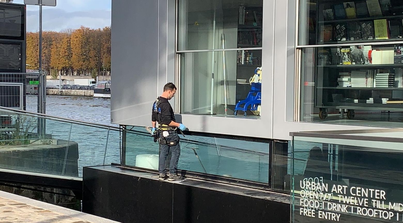 Un homme portant un harnais de sécurité nettoie les vitres d'un bâtiment en bord de rivière.