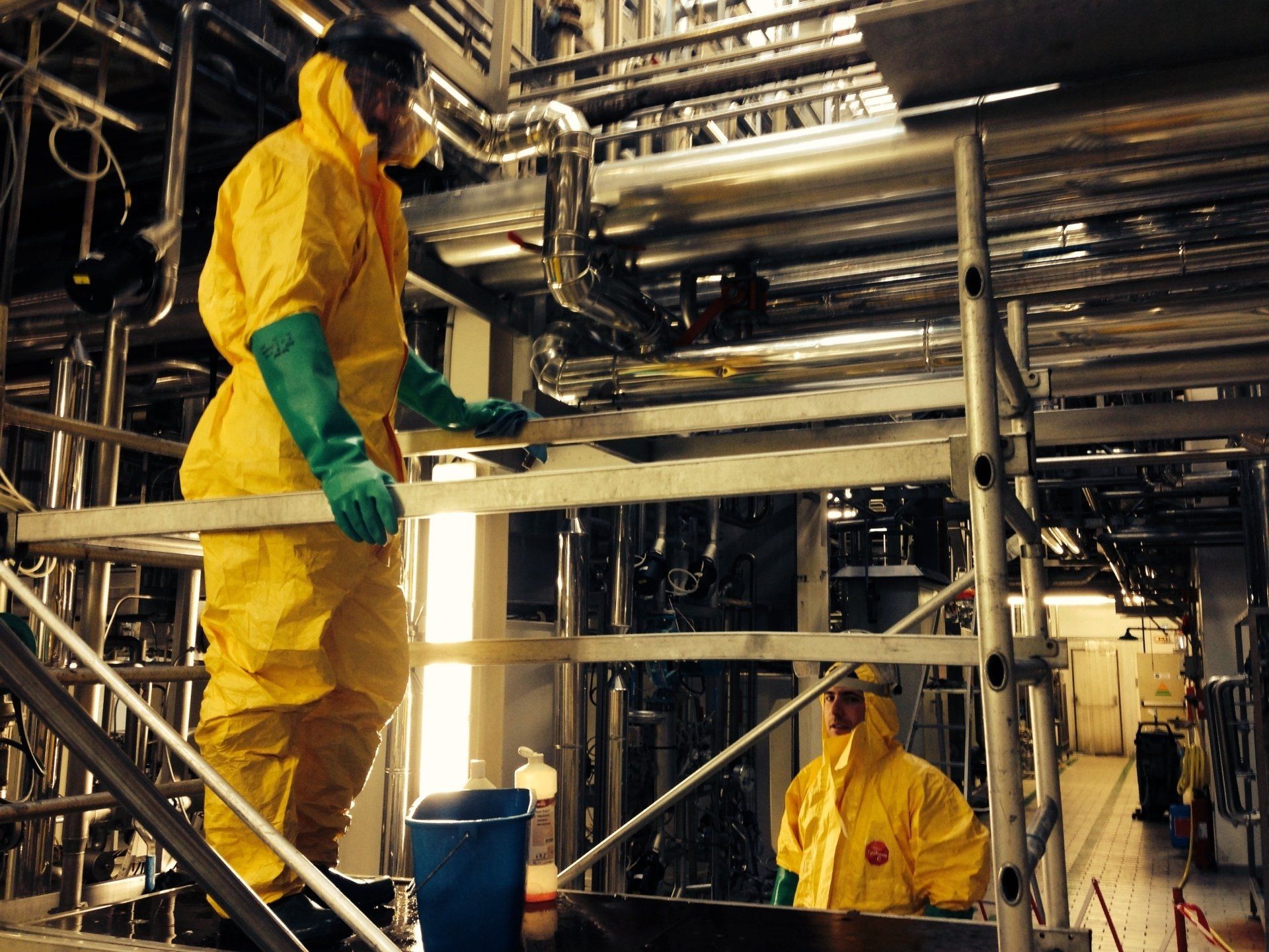 Deux personnes vêtues de combinaisons de protection jaunes inspectent des tuyaux métalliques.