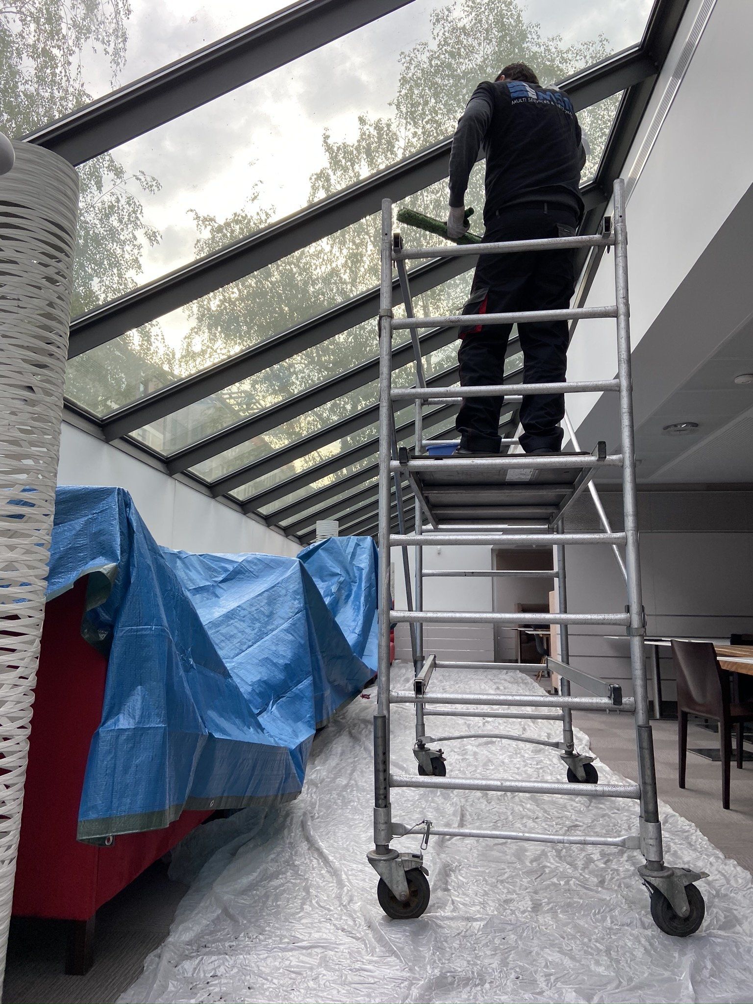 Une personne nettoie un plafond de verre sur une échelle roulante. Des meubles sont recouverts d'une bâche bleue. Intérieur.