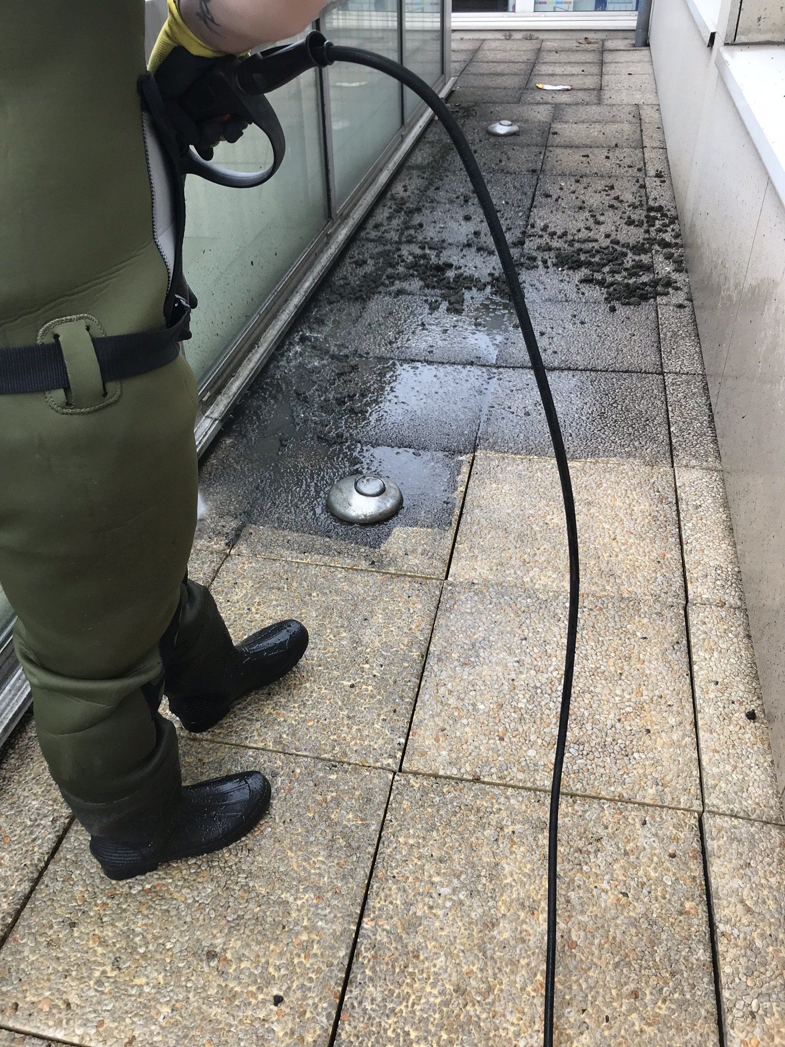 Une personne en cuissardes utilise un tuyau d'arrosage pour nettoyer une terrasse mouillée.