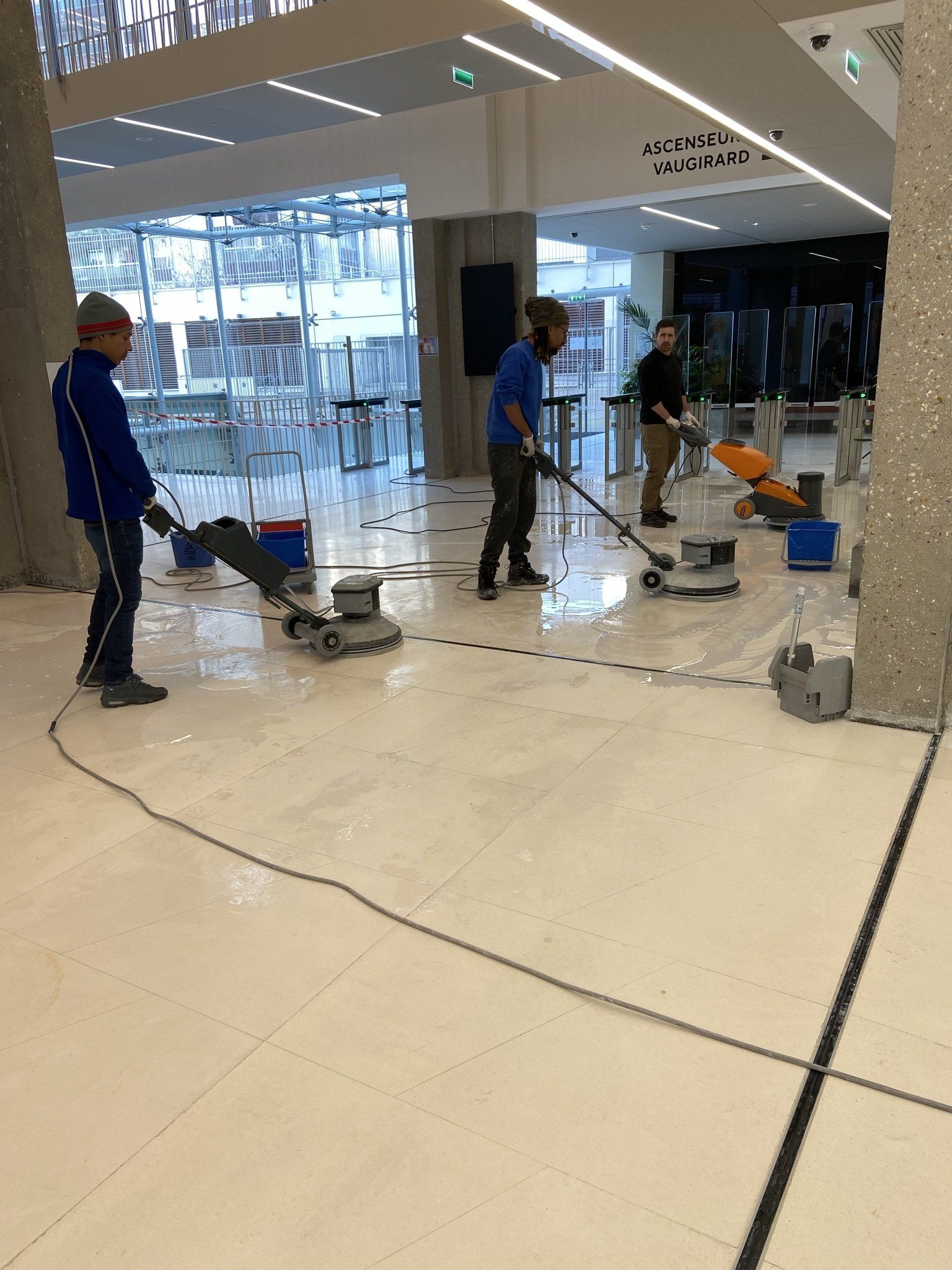 Trois personnes polissent un grand sol clair dans un immeuble moderne.