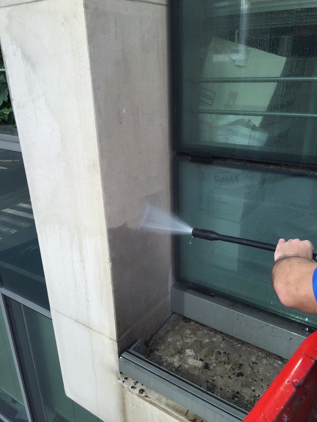 Une personne nettoie au nettoyeur haute pression la colonne en pierre extérieure d'un bâtiment, près d'une fenêtre.