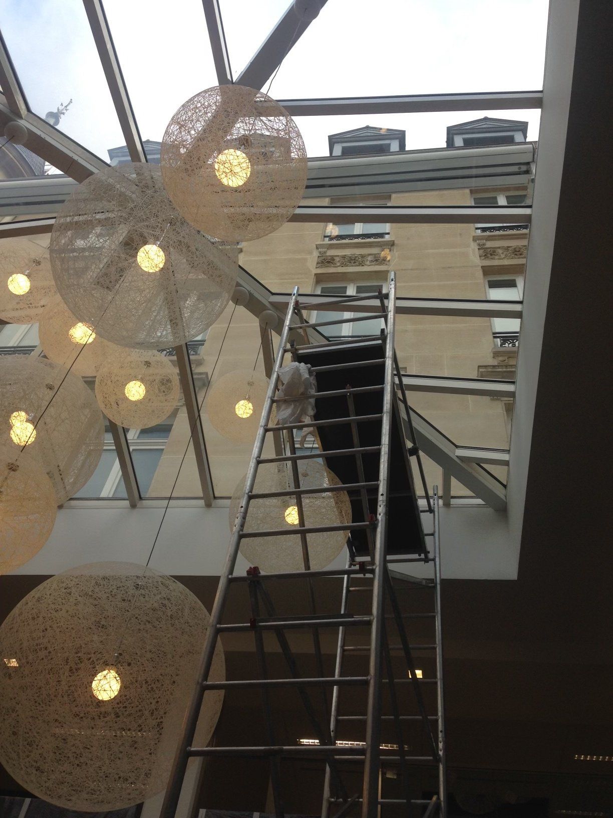 Une personne sur une échelle travaille sur des luminaires suspendus dans un bâtiment à toit de verre.