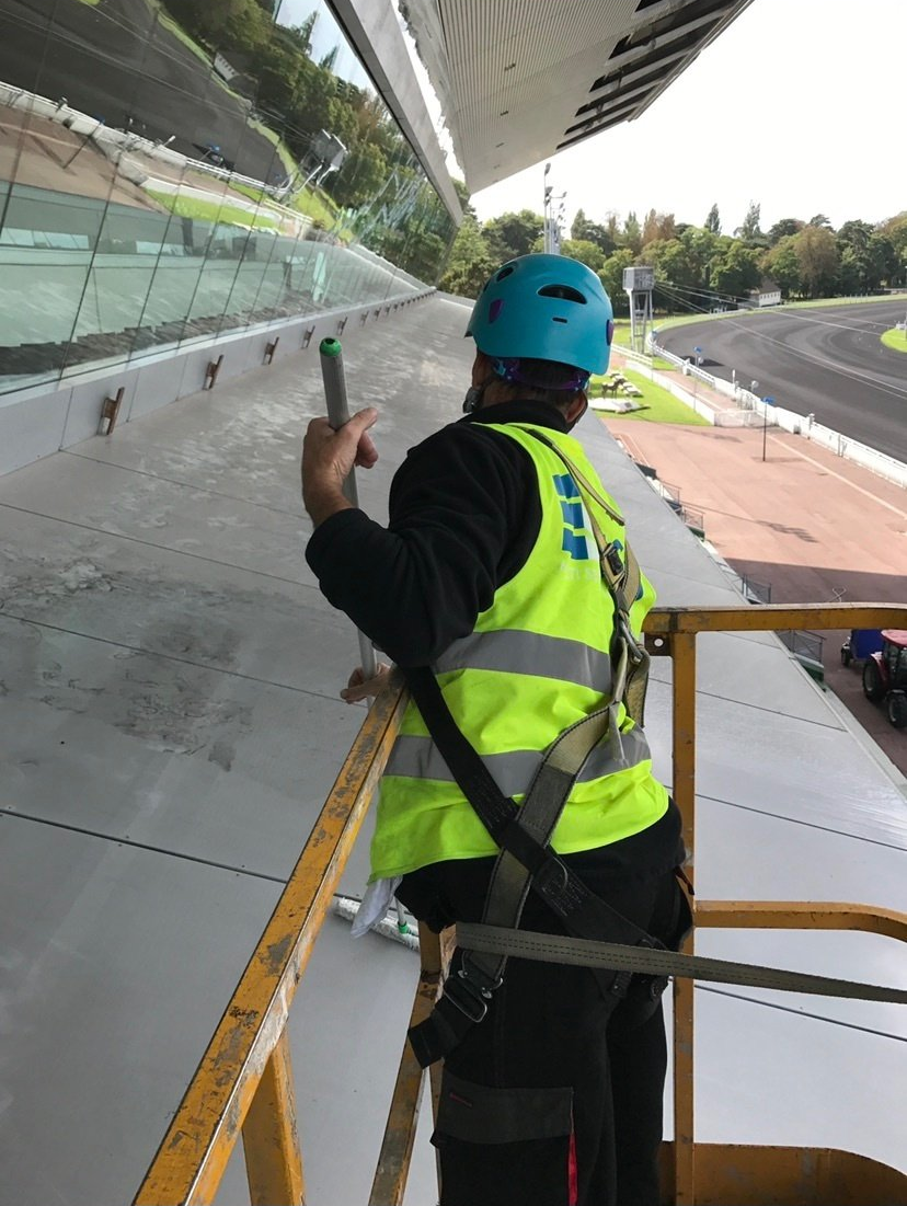 Personne en tenue de sécurité nettoyant une paroi vitrée donnant sur un circuit automobile.
