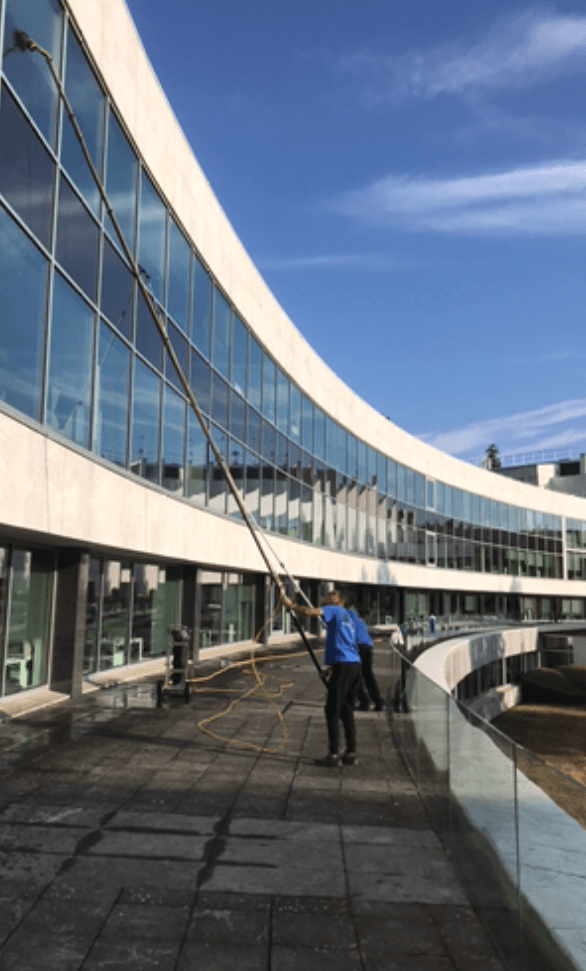 Par une journée ensoleillée, une personne lave les grandes fenêtres incurvées d'un immeuble à l'aide d'une longue perche.