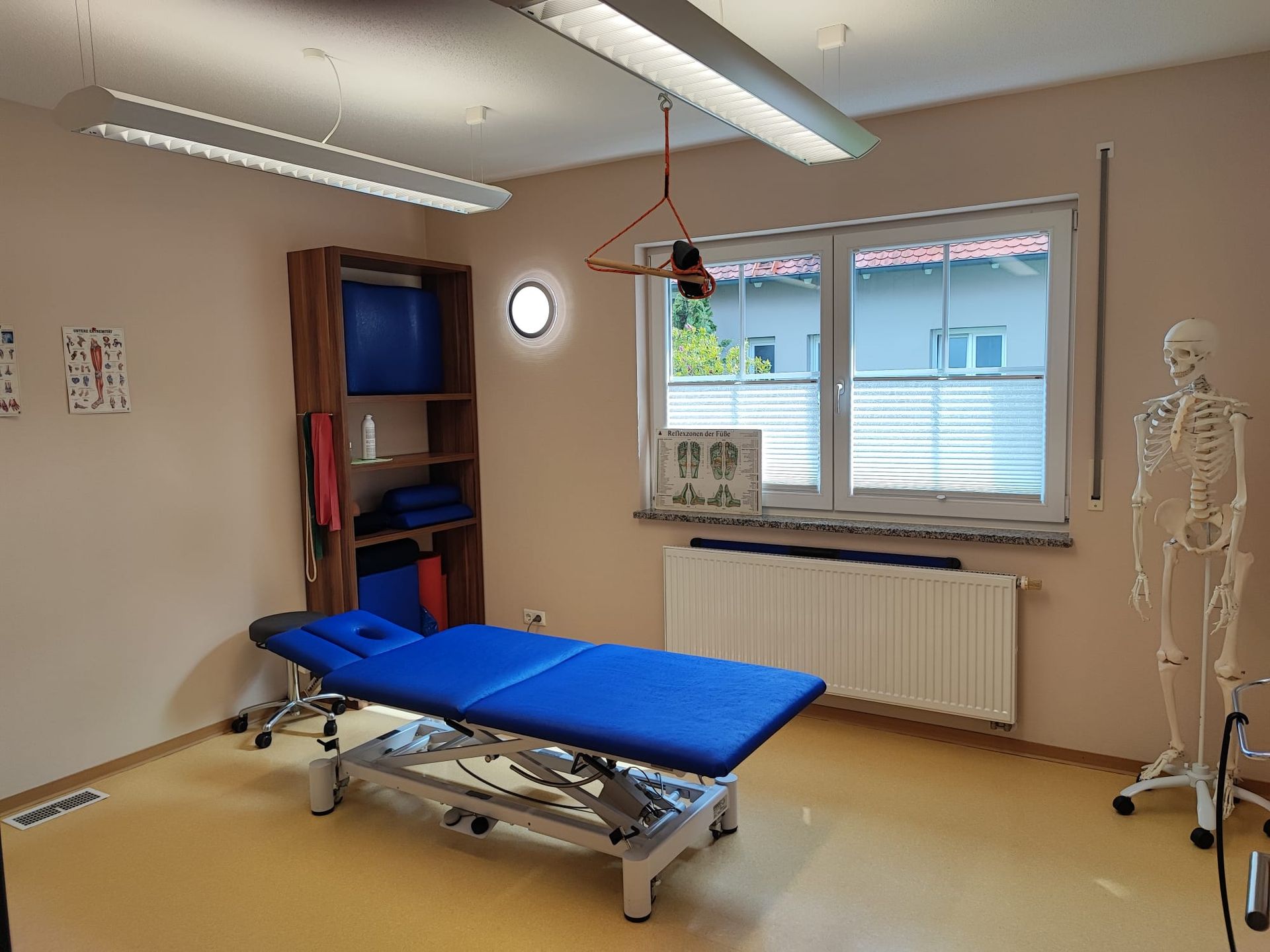 Therapieraum mit blauem Behandlungstisch, Regalen, Fenster und Skelettmodell.
