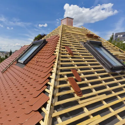 Toiture en cours de réalisation, pose de tuiles rouges sur une charpente en bois, avec puits de lumière et cheminée sur fond de ciel bleu.