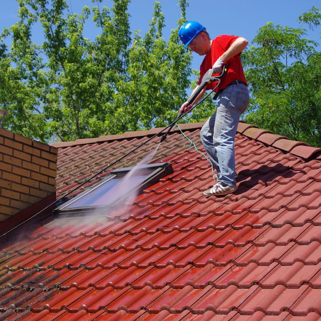 Une personne en chemise rouge et casque bleu nettoie au jet d'eau un toit de tuiles rouges.