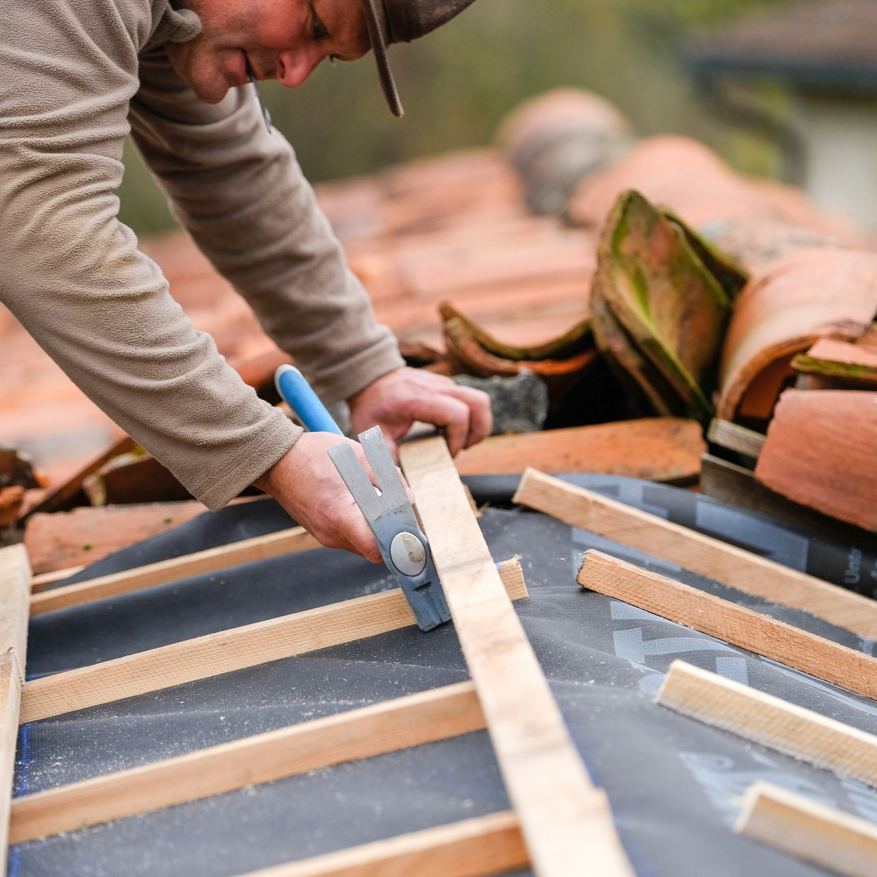 Un couvreur utilise un marteau pour fixer des lattes de bois sur un toit recouvert d'une sous-couche noire.