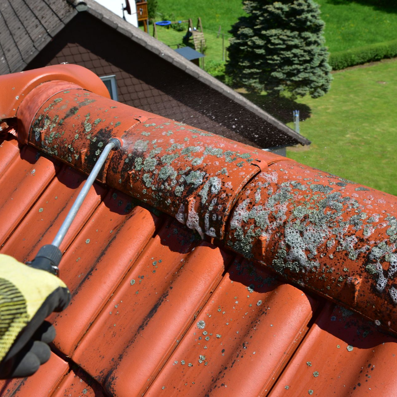 Une personne gantée arrosant au jet d'eau un toit de tuiles rouges sales.