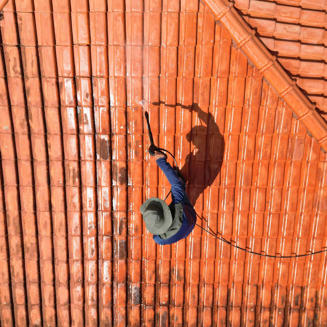 Une personne nettoie à haute pression un toit de tuiles orange.