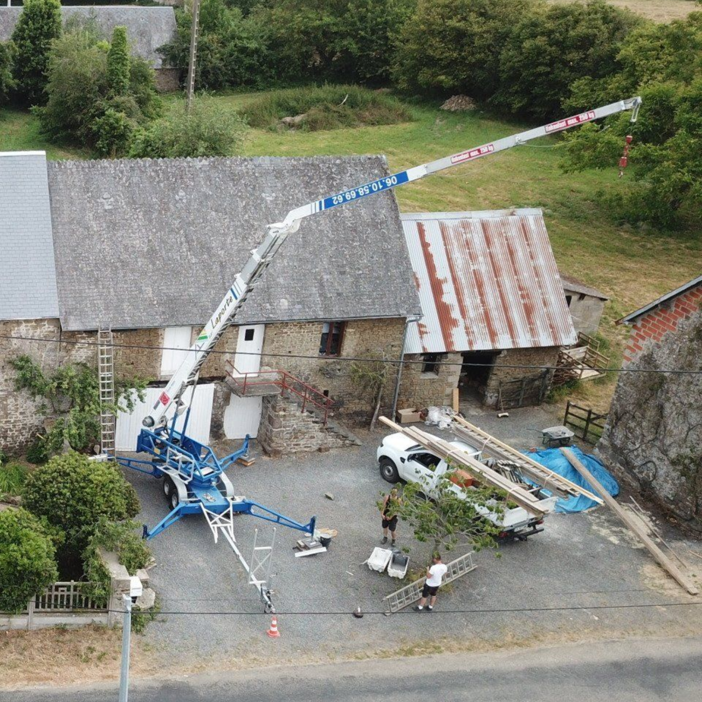 Une plateforme élévatrice bleue s'avance vers un bâtiment en pierre où se trouvent des ouvriers, du matériel et un camion blanc.
