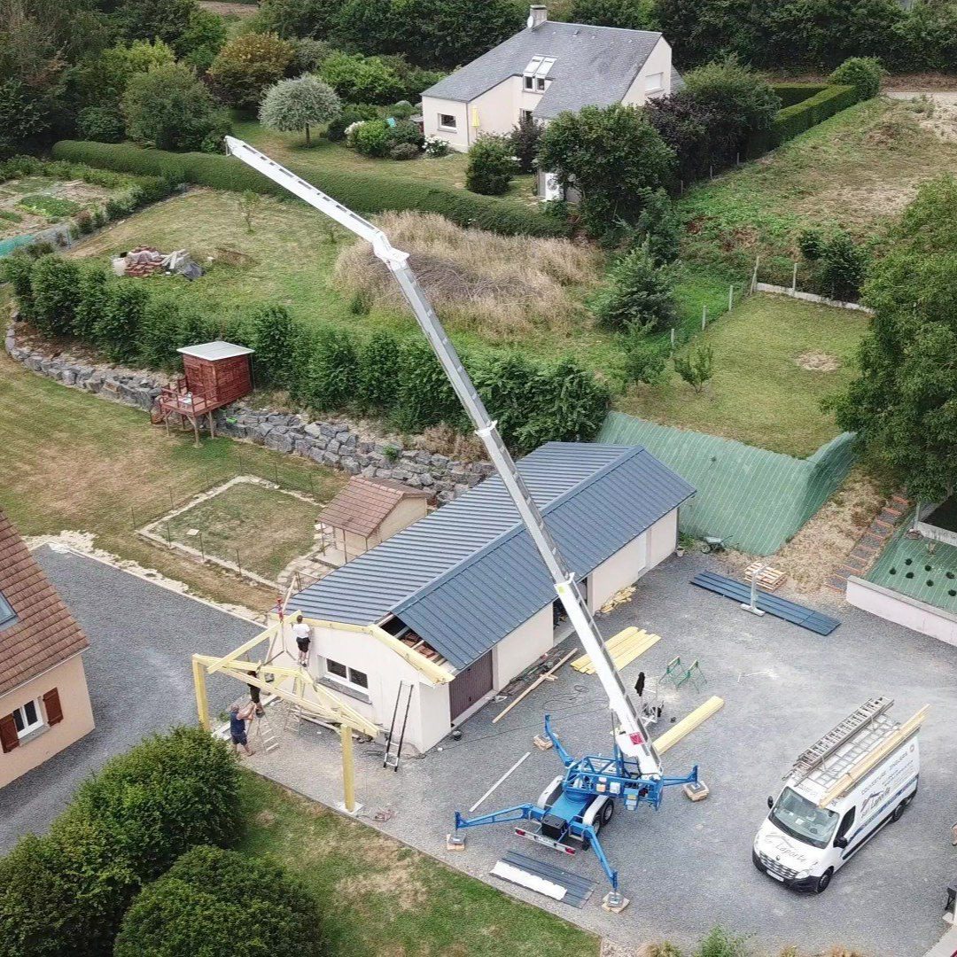 Chantier : ascenseur bleu surplombant un bâtiment au toit gris ; camionnette blanche et maisons en arrière-plan.