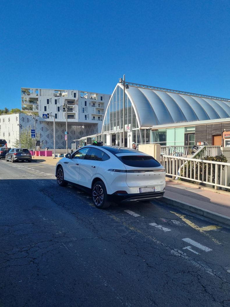 Un SUV blanc est garé dans une rue ensoleillée, à côté d'un bâtiment moderne au toit métallique incurvé.