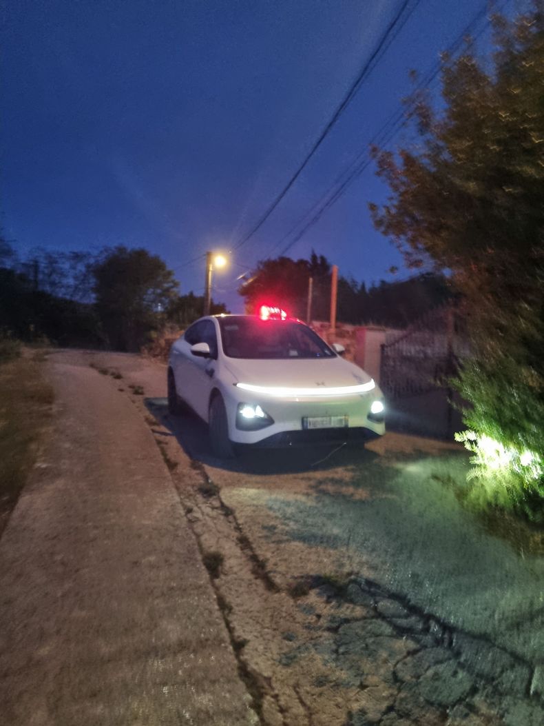 Voiture blanche garée sur le bas-côté d'une route défoncée au crépuscule, phares allumés et feux arrière rouges visibles.