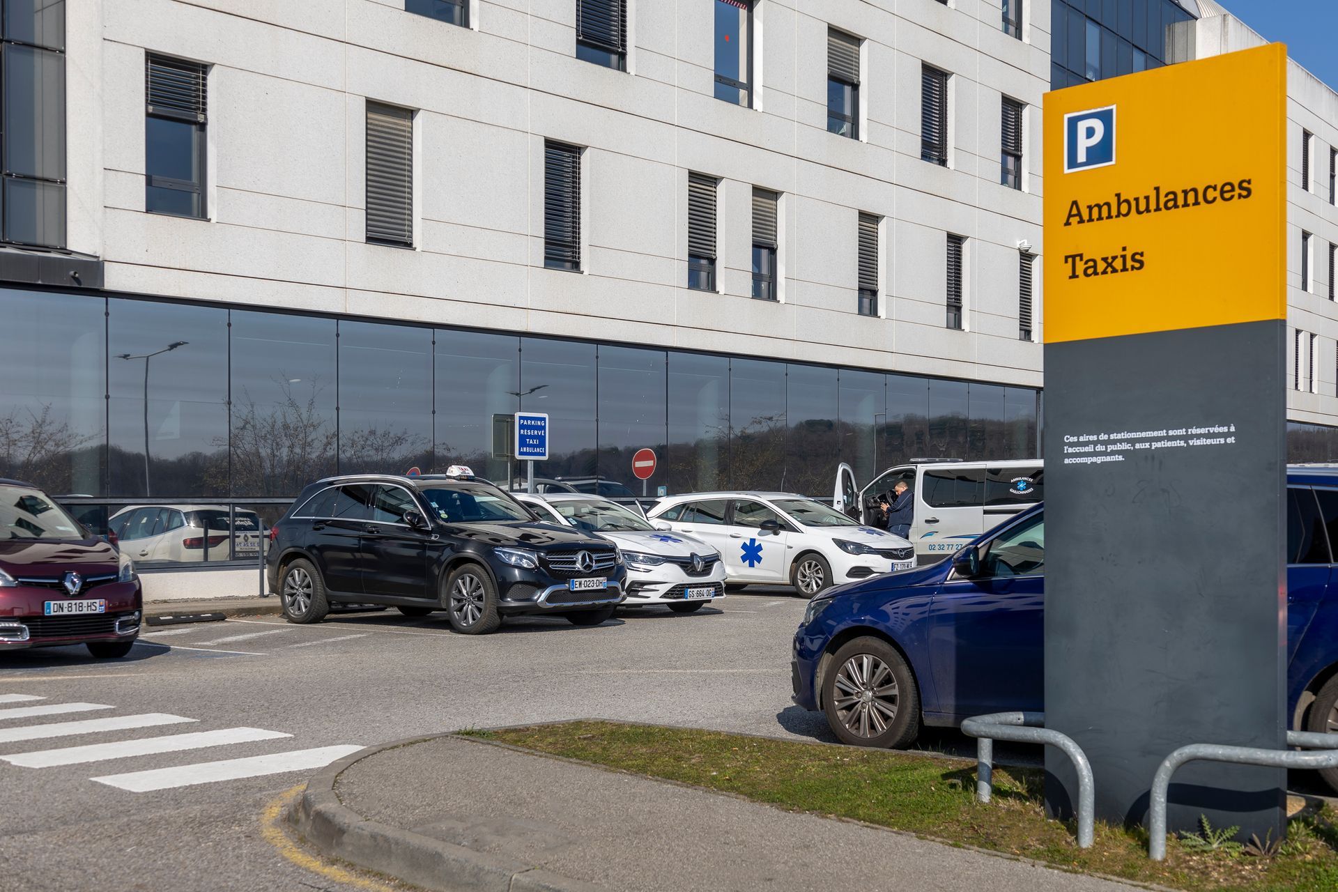 Entrée de l'hôpital avec voitures garées et un panneau indiquant les ambulances et les taxis.