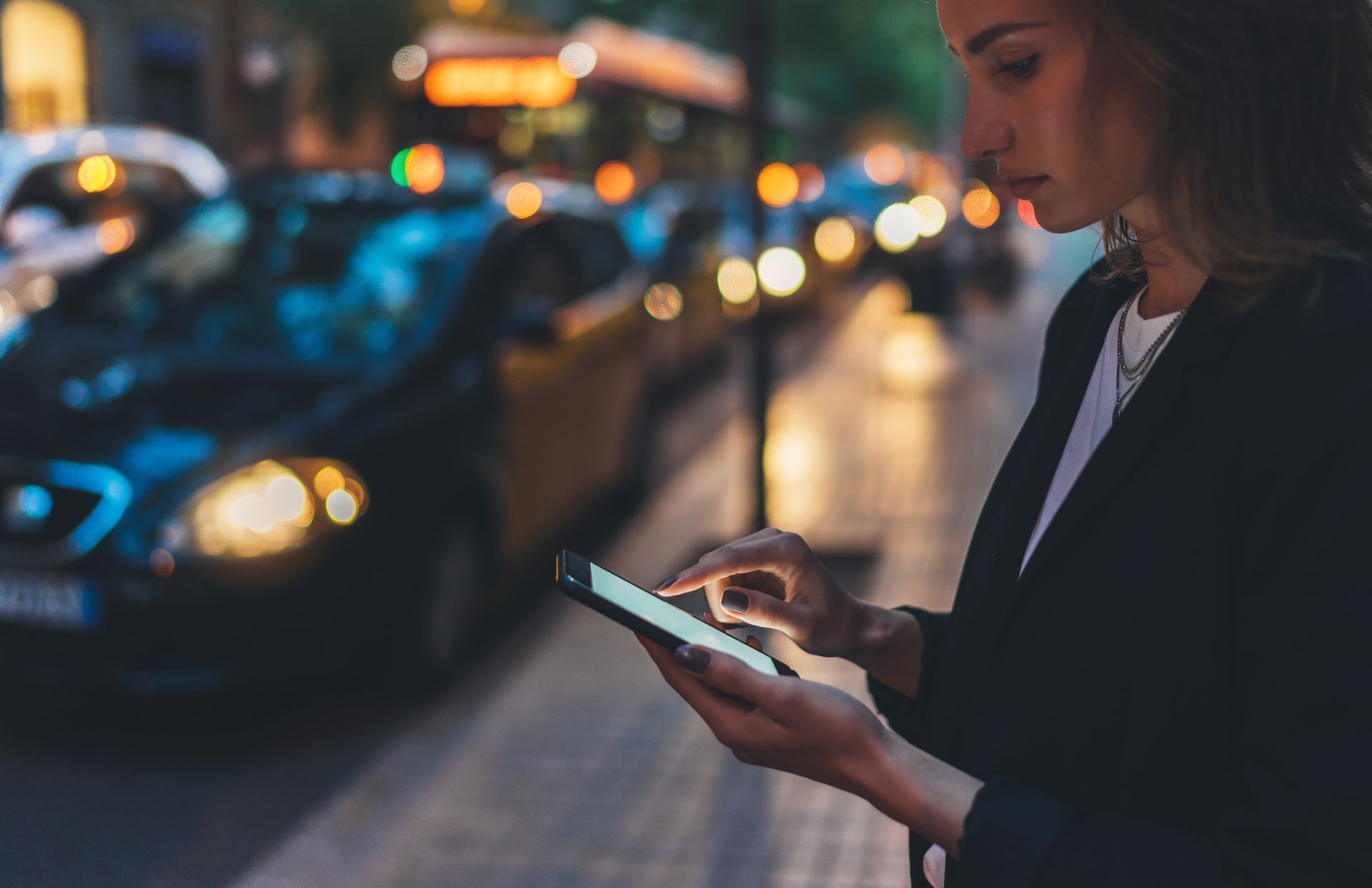 Une femme utilise un smartphone sur un trottoir en ville, la nuit, avec des phares de voiture flous en arrière-plan.