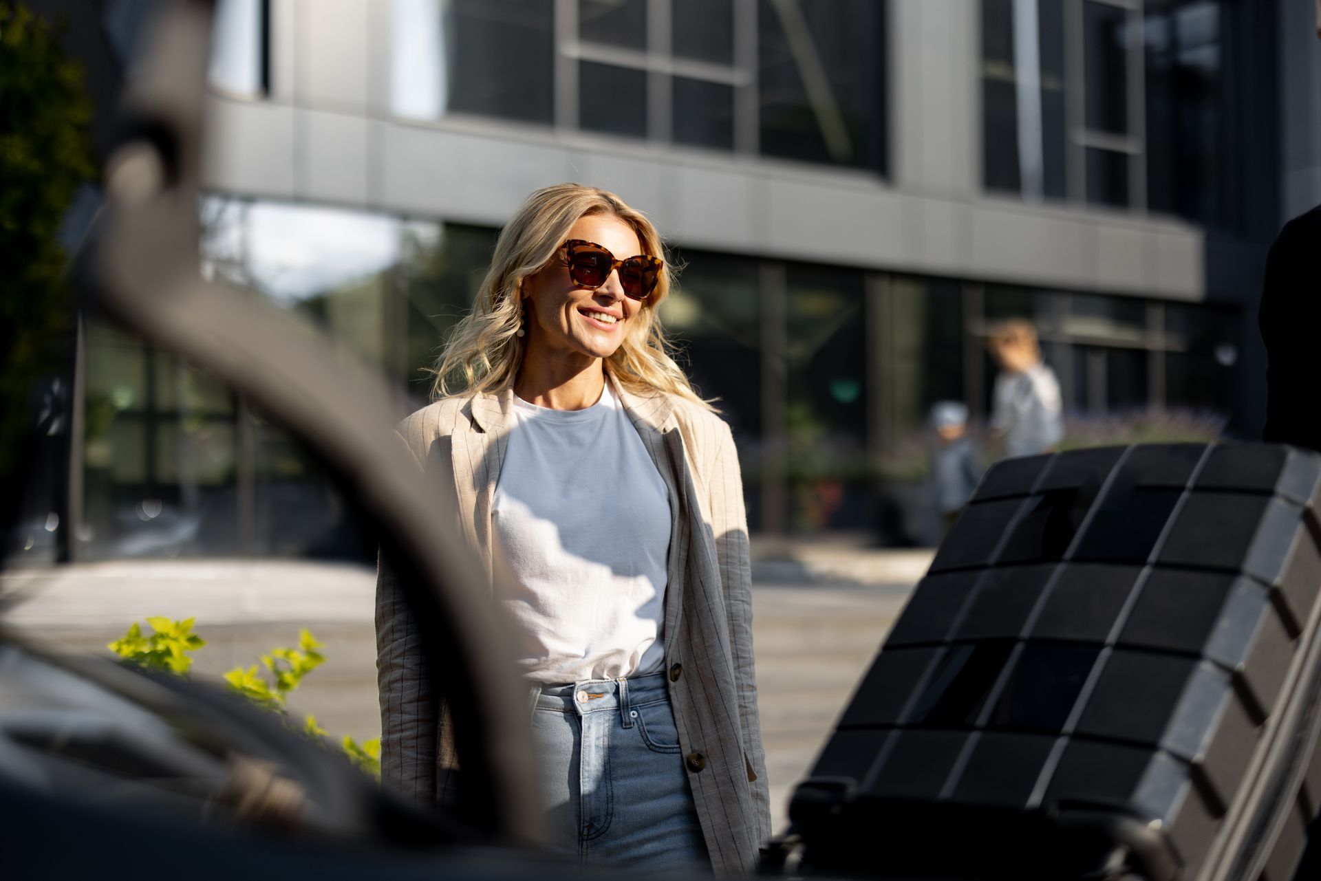 Une femme portant des lunettes de soleil se tient à l'extérieur, à côté d'une voiture noire, dans une rue ensoleillée.