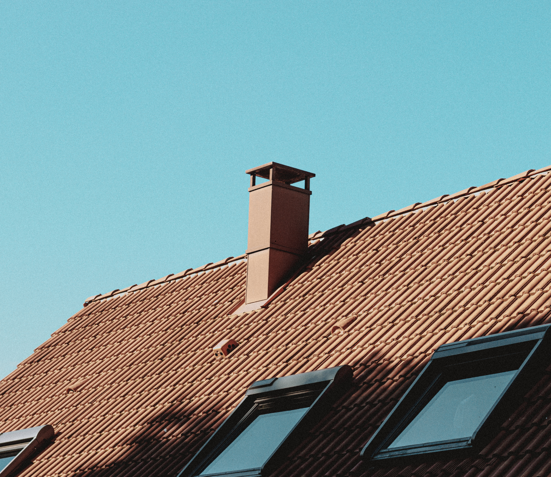Toit de tuiles rouges avec cheminée et puits de lumière contre un ciel bleu clair.
