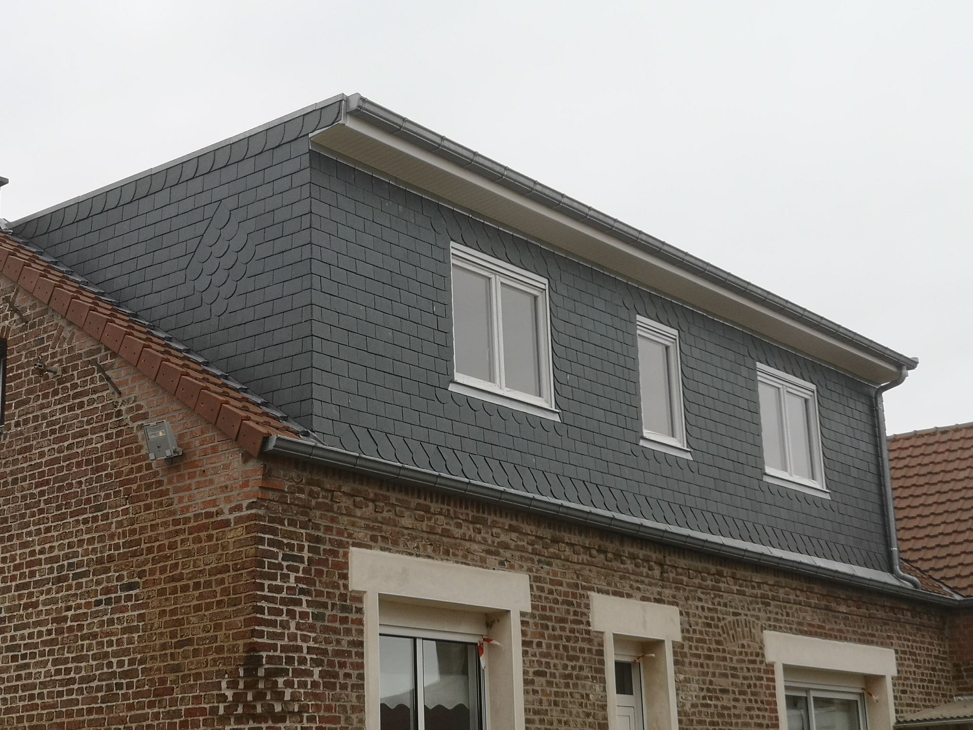 Bâtiment en briques avec une lucarne recouverte d'ardoise gris foncé et des fenêtres à cadre blanc.