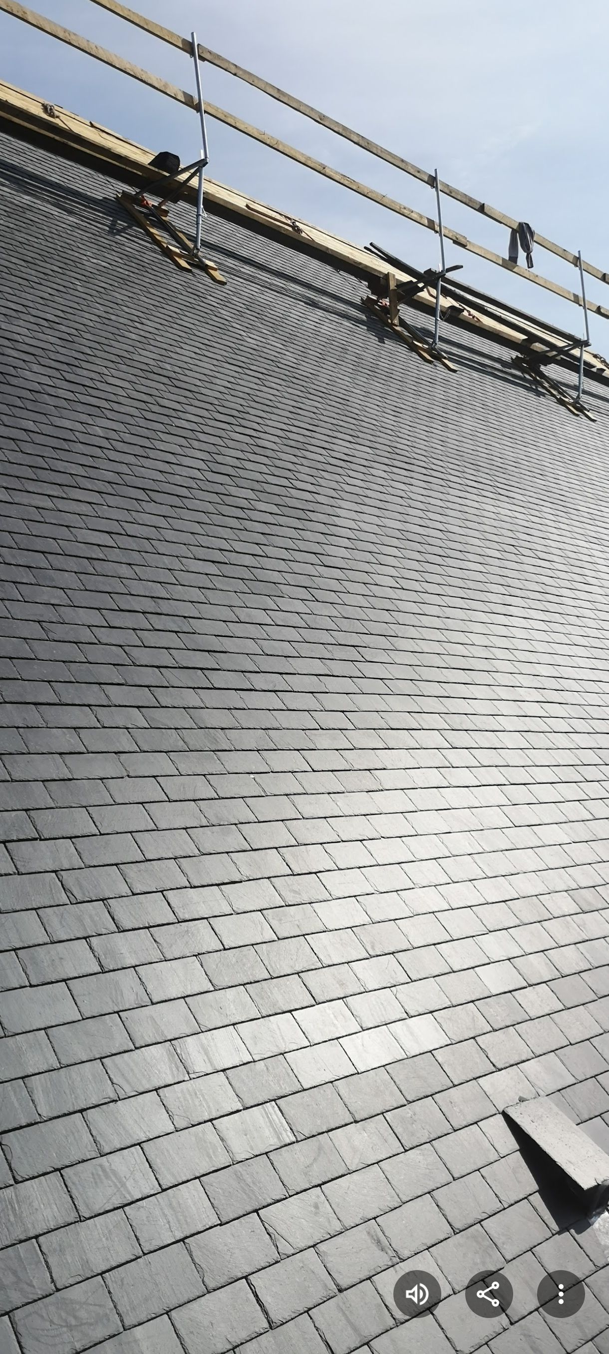 Un toit recouvert de tuiles d'ardoise gris foncé. Un échafaudage est positionné près du sommet, sur fond de ciel bleu clair.